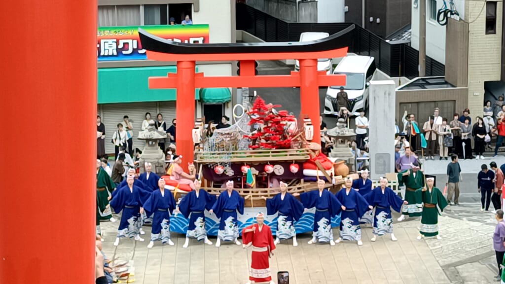 Men in blue costumes lined up in front of Aburaya-machi's kawafune float, beneath a red torii gate at Yasaka Shrine in Nagasaki, during Niizoroi