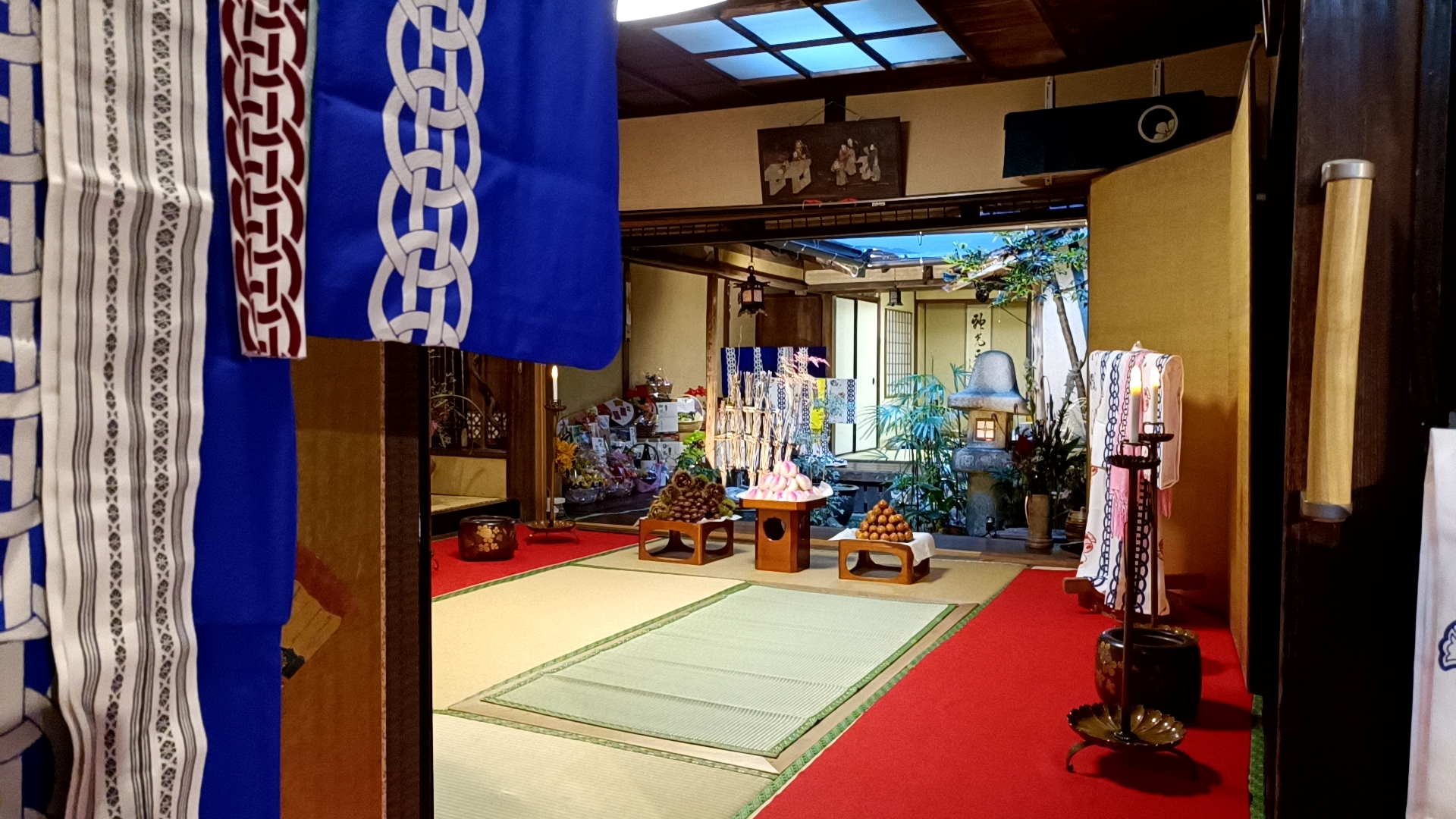 A tatami room with a red carpet, stacked offerings and a small inner garden with a stone lantern, seen during Niwamise in Kajiya-machi, Nagasaki