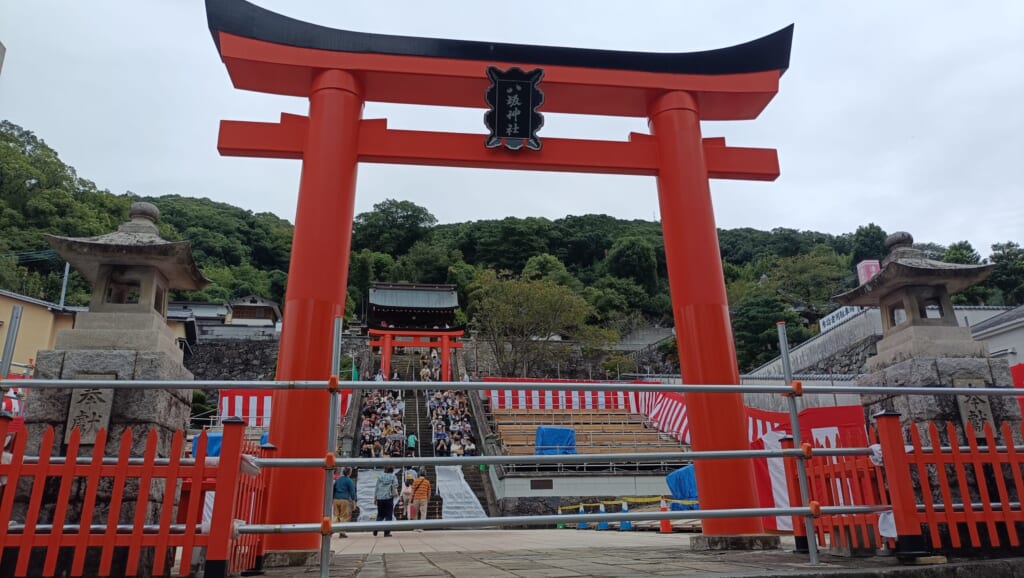 The red torii gate of Yasaka Shrine in Nagasaki, with spectators sitting on the stone steps beyond it and festival stands to the right