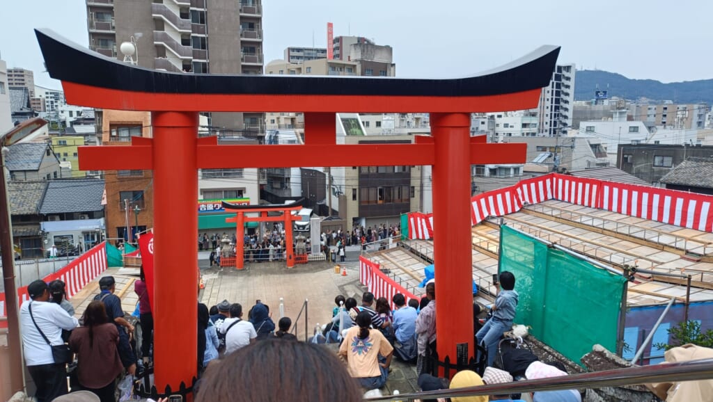 Spectators on the stone steps of Yasaka Shrine looking down through a torii gate at the open space where Niizoroi is performed