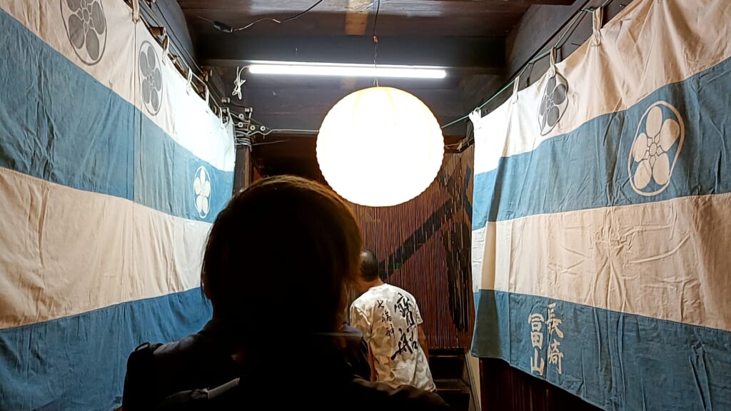 A narrow passage hung with faded blue-and-white curtains bearing a family crest, leading to a Niwamise display in Nagasaki