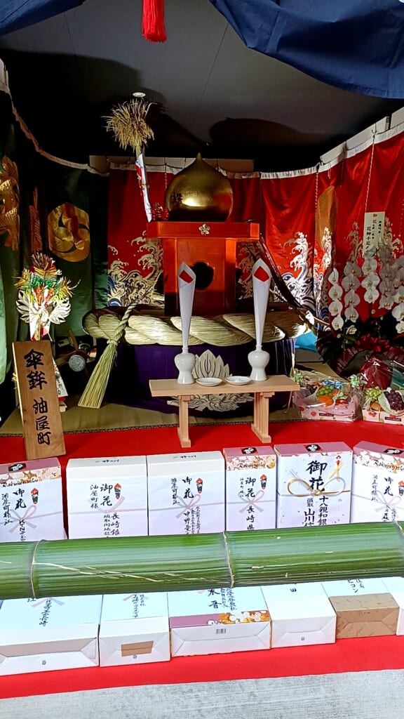 A golden ornament, a thick straw rope and embroidered hangings from Aburaya-machi's kasaboko, displayed above rows of gift boxes