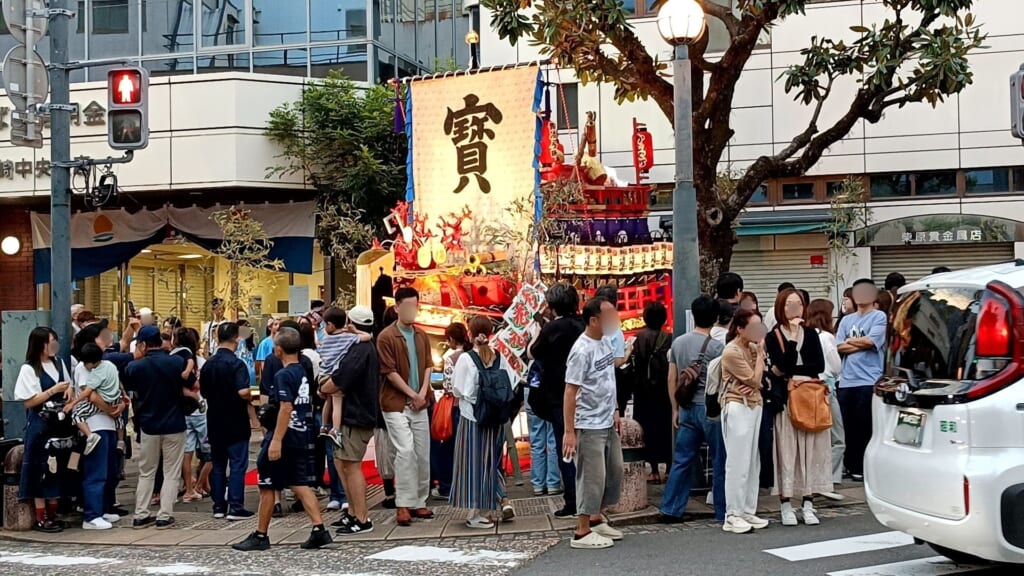 Kajiya-machi's treasure ship float, its sail bearing the character for treasure, surrounded by a crowd at a street corner in Nagasaki